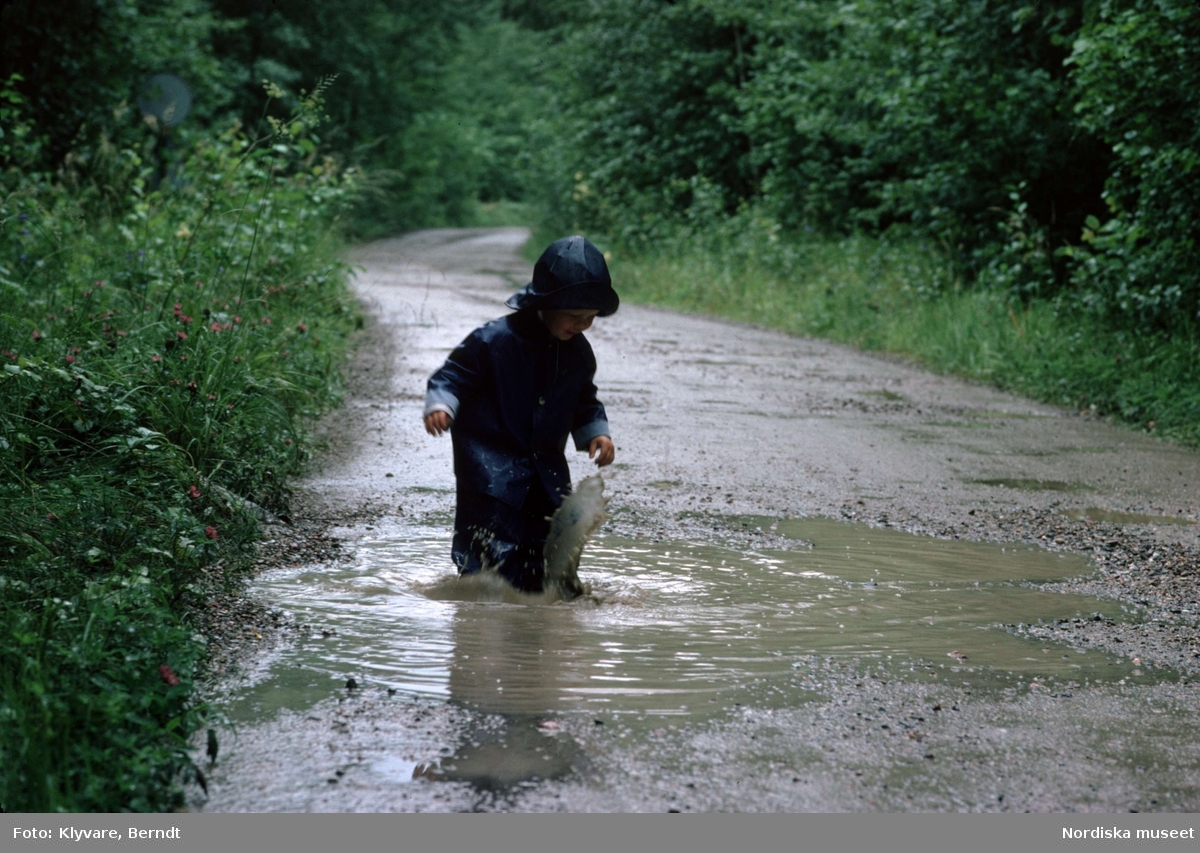 Ett litet barn i galonkläder plaskar i en vattenpöl på en regnvåt väg.