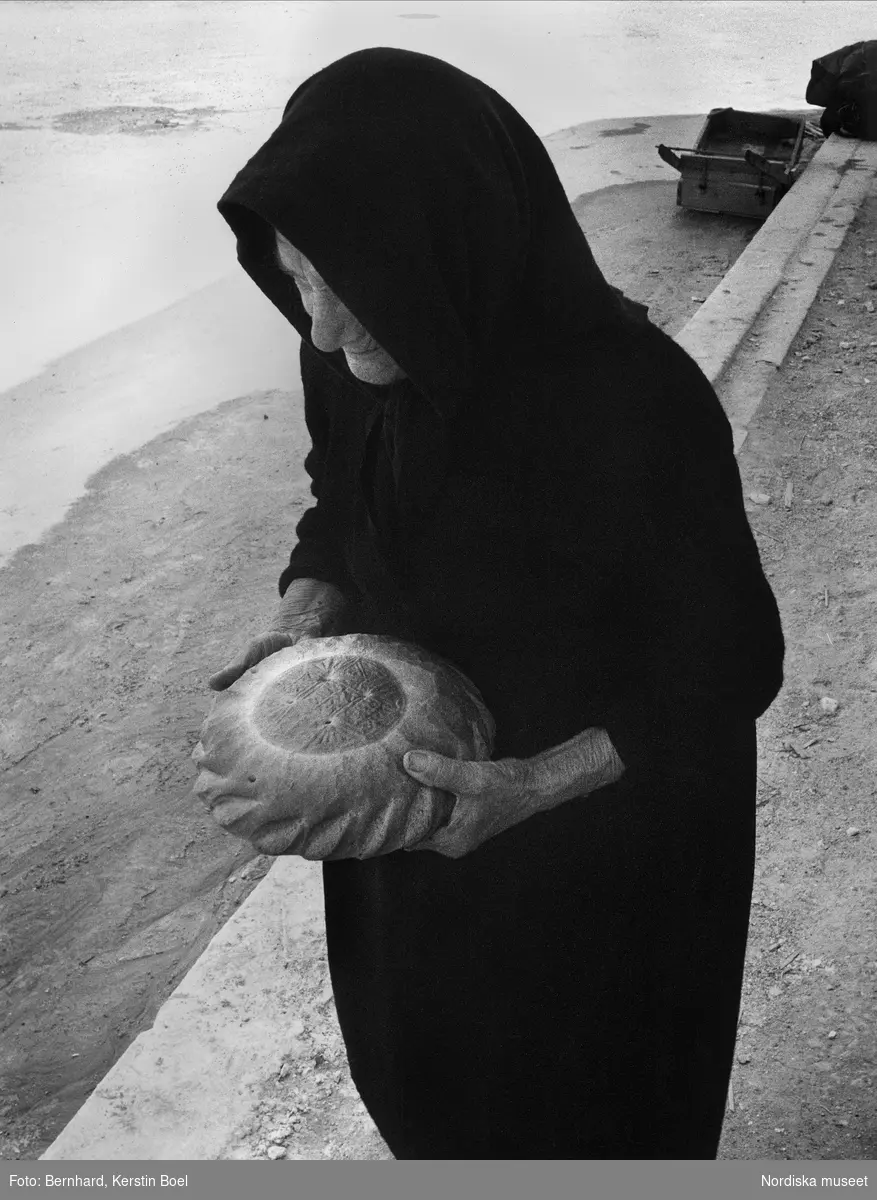 Påskbröd. En äldre kvinna i svarta kläder bär ett runt nattvardsbröd, Lebadeia, Grekland. Brödet är stämplat med religiösa symboler och bakas till den ortodoxa kyrkans påskfirande. Det skärs i små kuber och doppas i vinet.