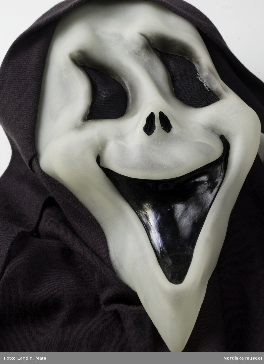 Liemansmask, halloweenutklädsel för barn, ansiktsmask av halvgenomskinlig vit plast med svart mun, ögonhål med svart nät, infälld i lång huva av svart bomullstrikå, hängande flikar vid öronen. Nordiska museets föremål inv.nr 328022.