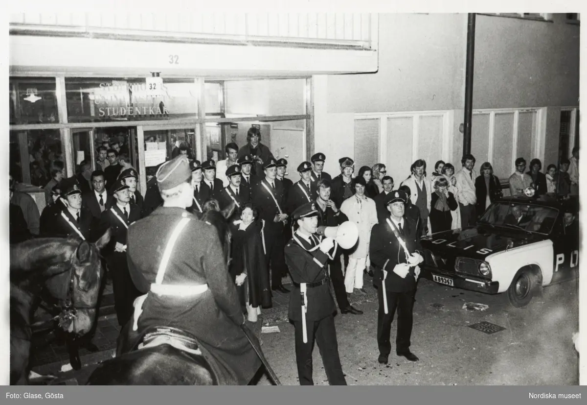 Kårhusockupationen vid Stockholms universitet i slutet av maj 1968. Kvällsbild från Studentkårens entré. Utanför har poliser barrikaderat dörrarna. En polisman talar till folksamlingen i megafon. En polisbil står parkerad intill. I förgrunden en ridande polis till häst.
