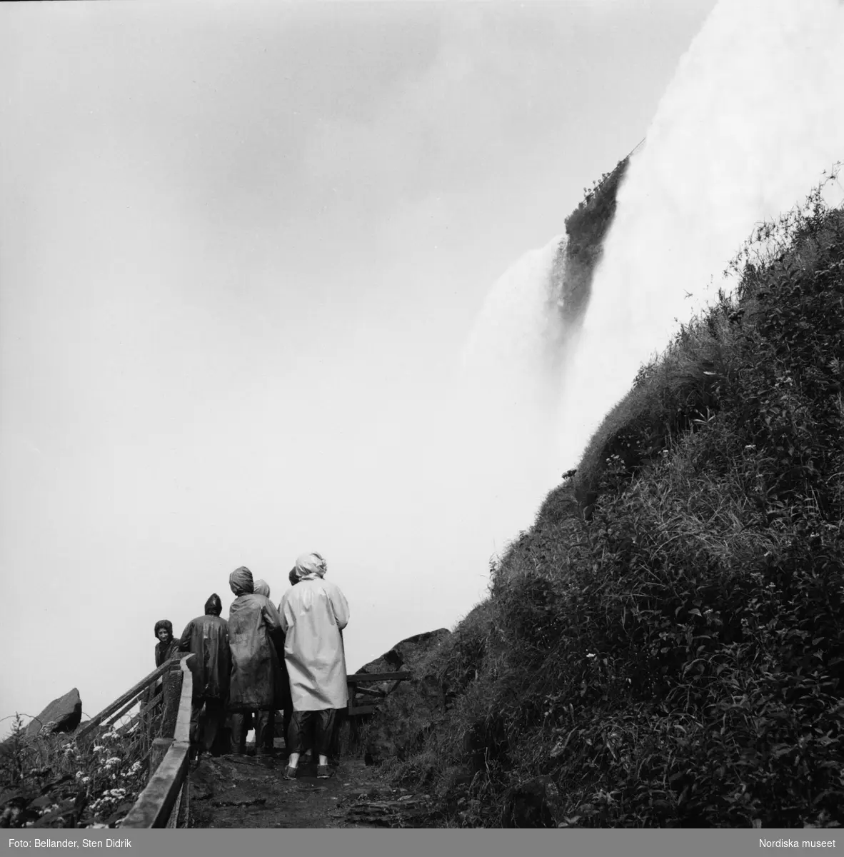 Regnklädda människor står vid Niagarafallet.
En grupp tittar upp mot det en annan står vid ett räcke och ser ner.