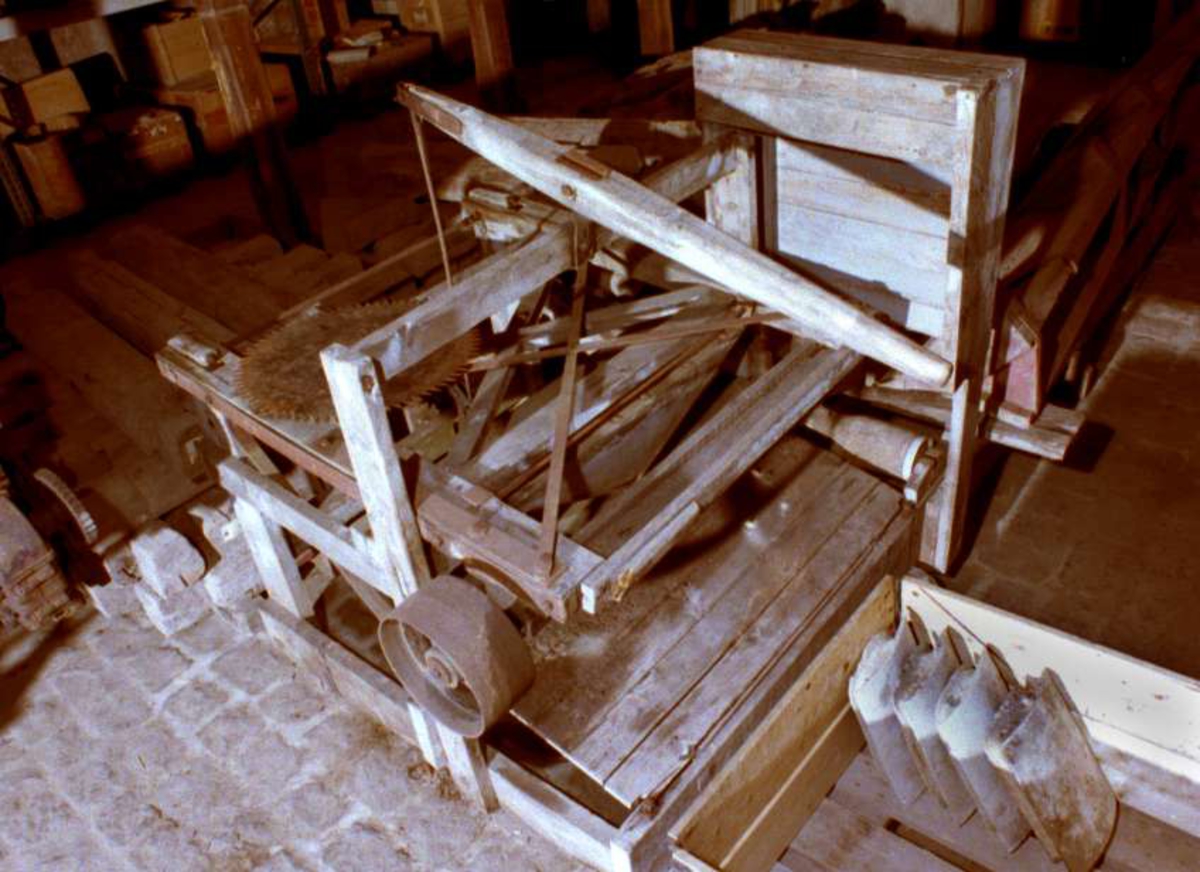Långvedkap för remdrift. Järnförstärkt träkonstruktion med en bana med fem trärullar, lådformigt skärbord med en lyftarm av trä och järn, och en fyrkantig lådformad uppbyggnad över sågklingan av järn som är lös, axel av järn med remhjul av järn.