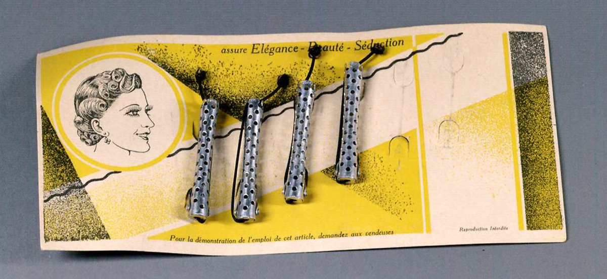 Fyra hårrullar av metall, perforerade och med tillhörande svart gummiband. Rullarna sitter på originalförpackningen i papp, med svart och gult tryck samt text på franska.