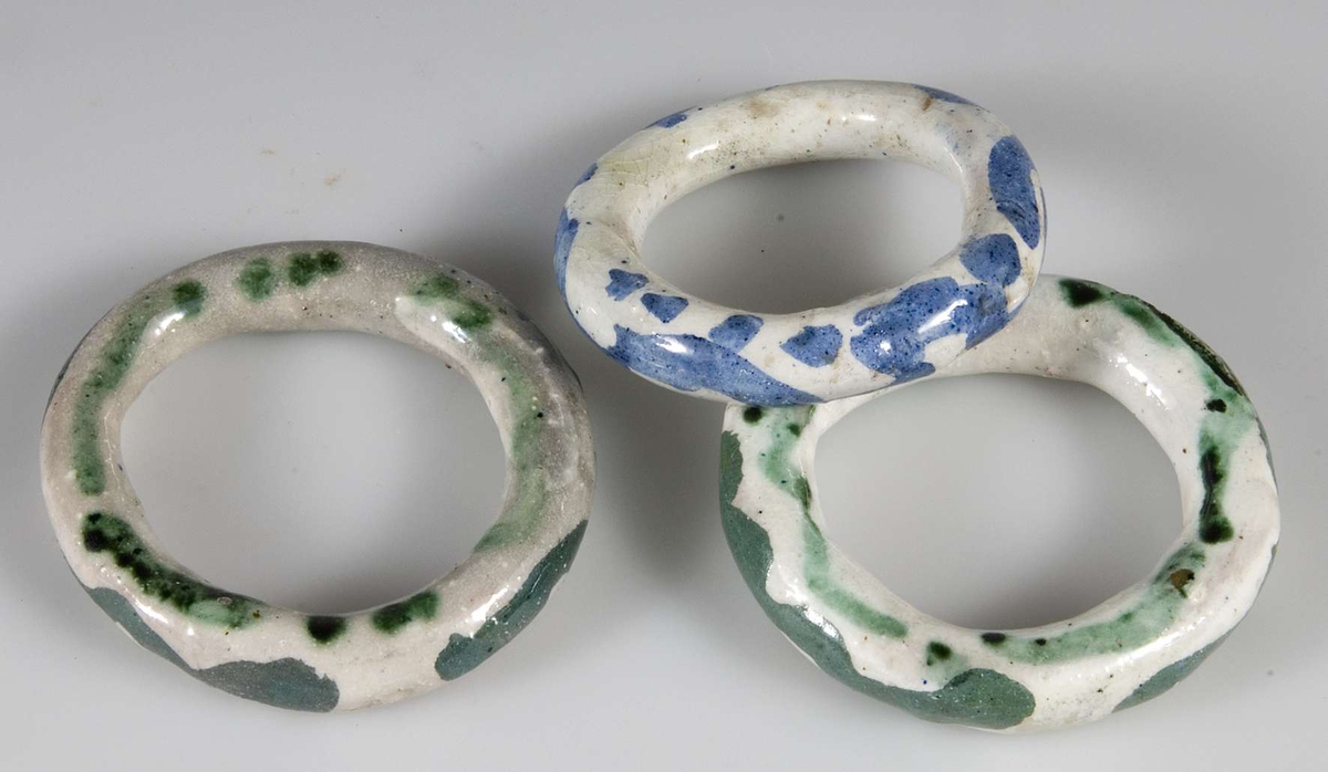 Tre gardinstångsringar av vitglaserat lergods. Två ringar med grönt mönster, en med blått.
