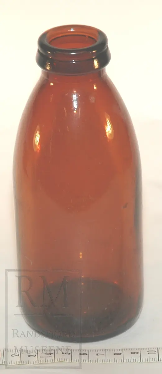 Sylinderformet flaske i brunt glass. Smalner inn mot toppen. Mangler kork.
