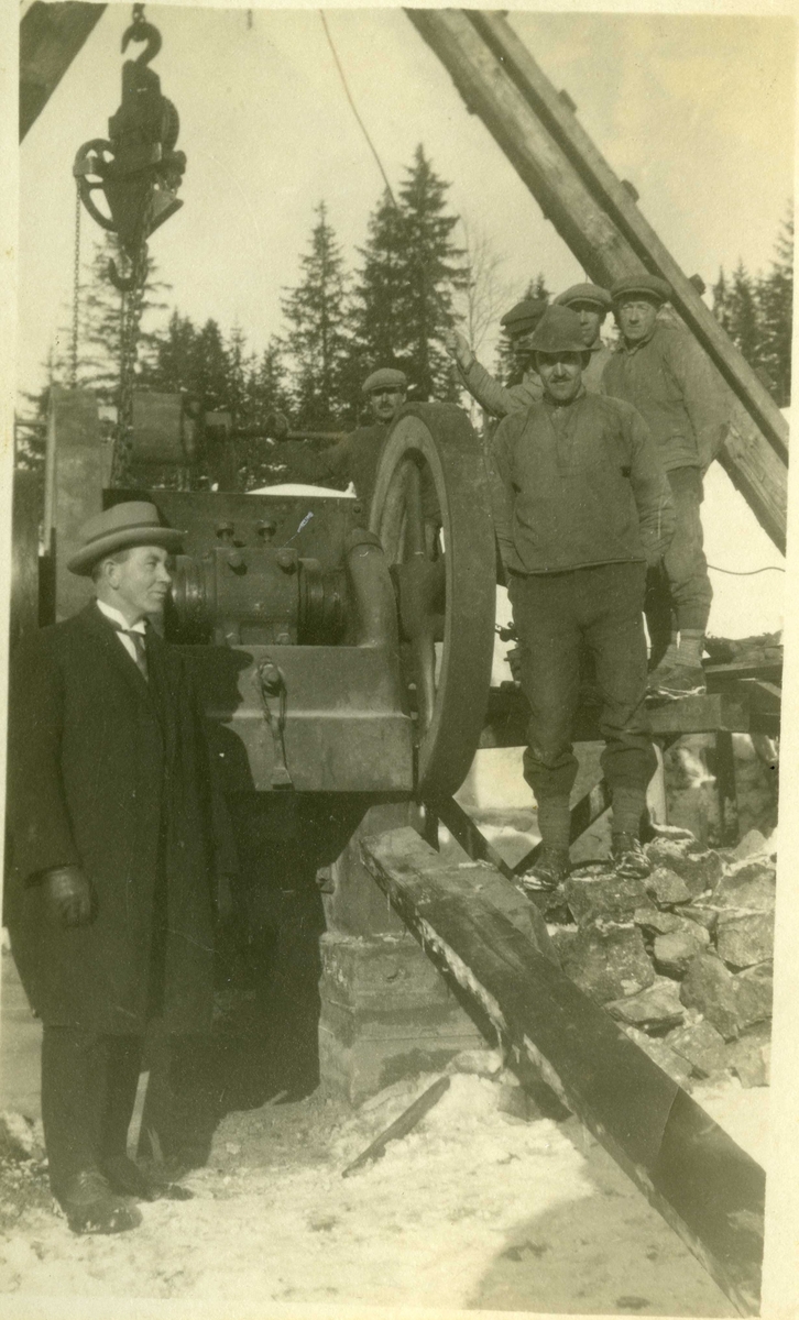 Fem arbeidere og en mann i dress og slips står ved en motor med drivverk