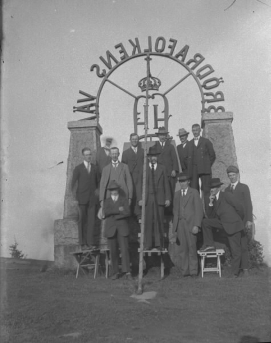 En grupp män på baksidan av kung Oscar II:s monument på Tabergs topp i Småland. Kungens valspråk "Brödrafolkets väl" bildar en båge mellan kraftiga naturstenspelare på ett fundament. Männen har klivit upp på monumentet och några sitter på bänkar nedanför. 

Monumentet tillkom 1895 i samband med att Oskar II invigde järnvägen mellan Nässjö-Jönköping, via Taberg, anlagd av Halmstad-Nässjö Järnvägs AB (HNJ). Efter att ha skrivit sitt namn på en berghäll nära stationen for konungen upp till bergets topp och därefter till Norrahammars bruk. Där spelade brukets musikkår folksången medan brukspatron J W Spånberg eskorterade majestätet in till gjuteriet där hans namnchiffer omgifvet av ett "Brödrafolkens väl" göts i sanden. HNJ lät så småningom utforma detta monument med det gjutna namnchiffret.
(Se även bild HB663)