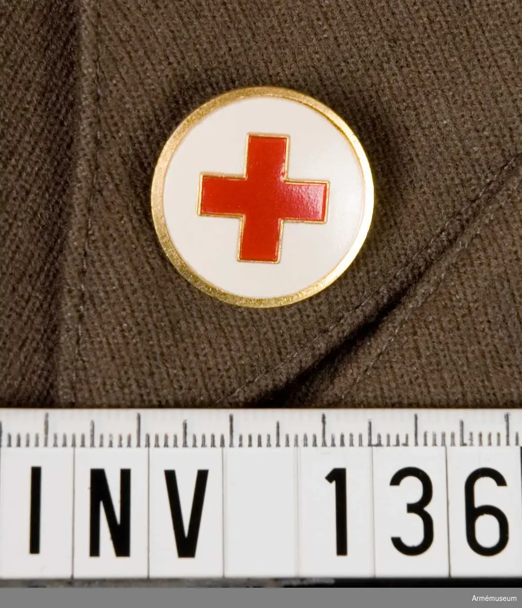 Samhörande nr är 132-144.
Märke m/1939-1953?, SRK.
Märkets framsida i mässing med vitmålad botten med ett rött kors. Baksidan har två runda hål avsedda för fastsättning.