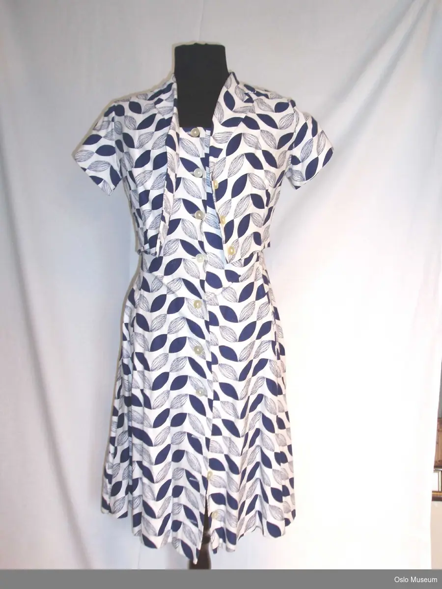Ermeløs kjole og kortermet bolero i hvit bunnfarge med mørkeblått bladliknende mønster. Knepping i hele kjolens lengde. To knapper i overdel.