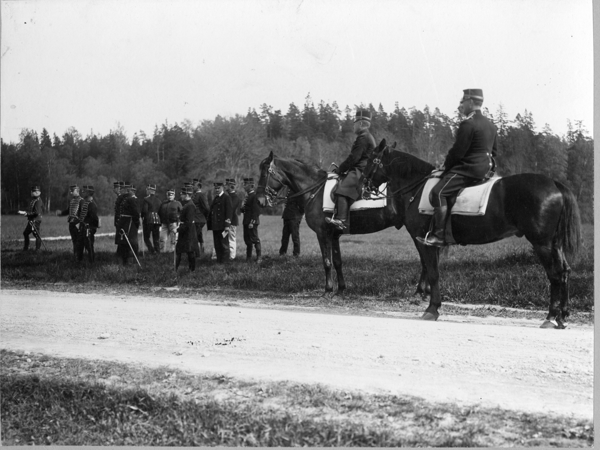 Två officerare till häst, tre husarer och ett antal officerare varav en sjöofficer står och väntar på något längs med landsväg.