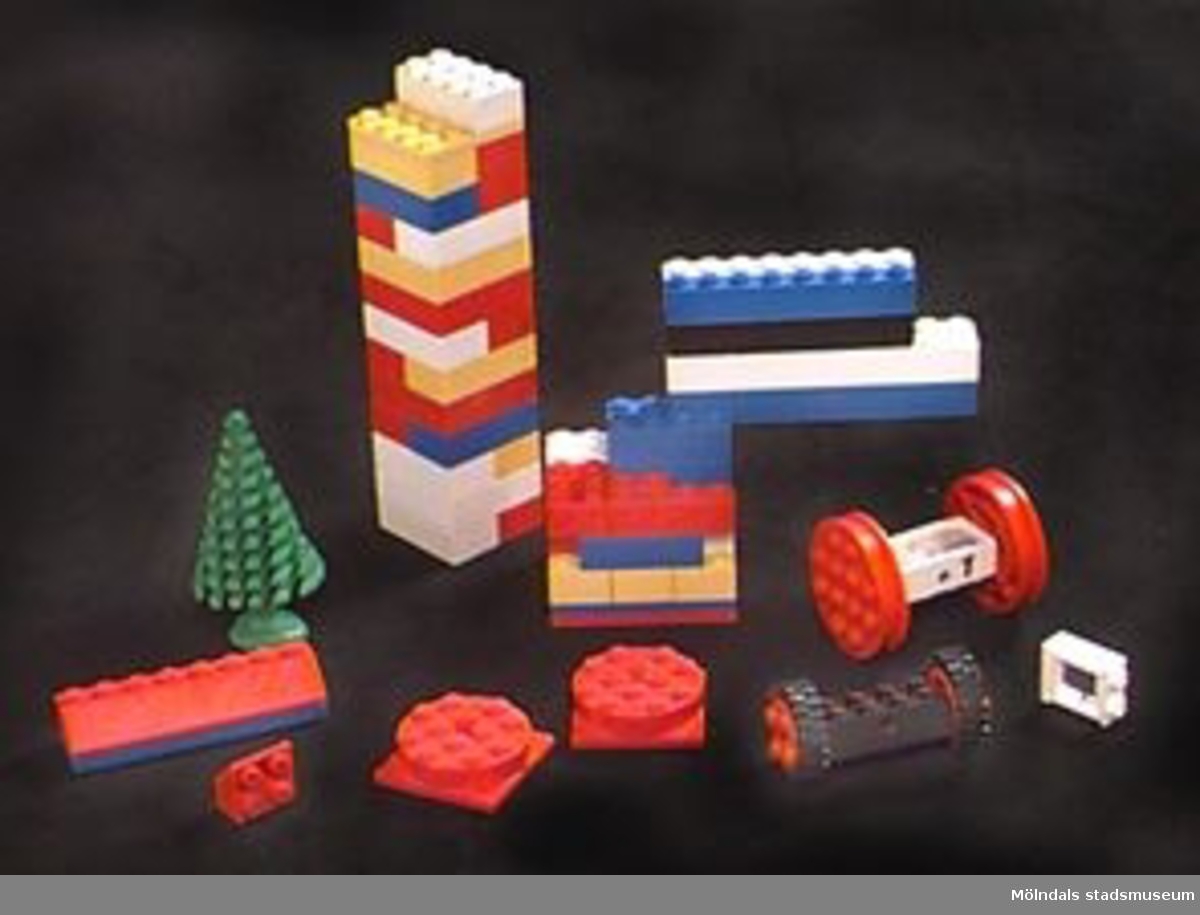Ett sjuttiotal legobitar varav en del är sammanfogade till mindre byggelement - 11 st allt som allt (se bilden). Förutom de vanliga rektangulära bitarna av olika storlek finns även: en grön gran, 2 hjulpar, 2 runda snurrbara socklar samt ett litet fönster. Bitarna förvaras i röd plastask. Mått: H 100mm. L 200mm. B 150mm.Katrinebergs daghem var kollektivt.Tidigare sakord: Legobitar.