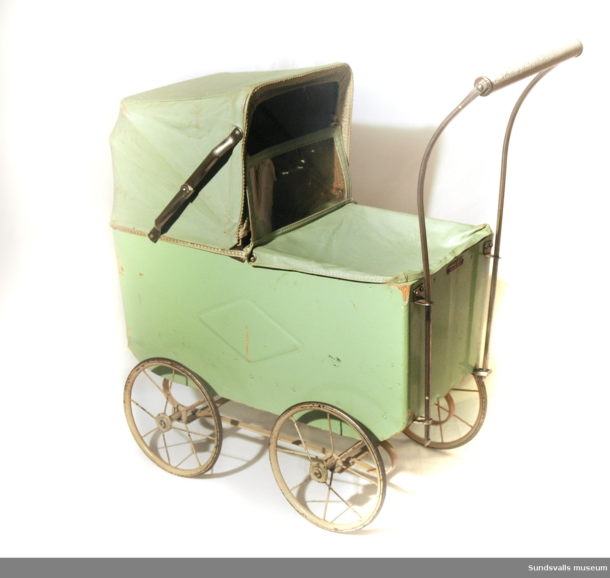 Dockvagn i ljusgrönt, med vagn av trä och suflett av vaxduk. På vagnens sida finns ett mönster i form av en fyrkant. I vagnen ligger även en virkad filt.