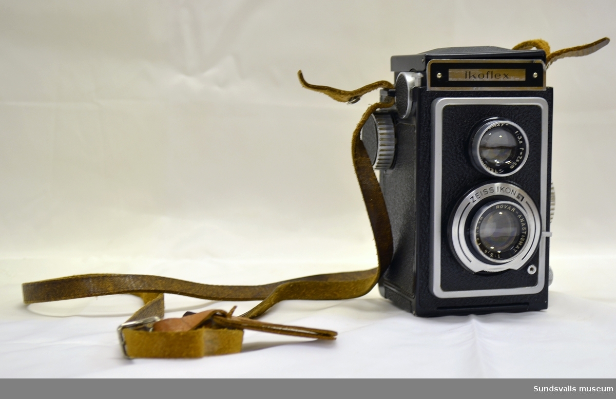 Svart kamera i metall, glas och plast med modellnamnet Iko Flex, från producenten Zeiss Ikon. Fastmonterad bärräm i brunt läder, varpå tidigare ägares namn står uppgivet.