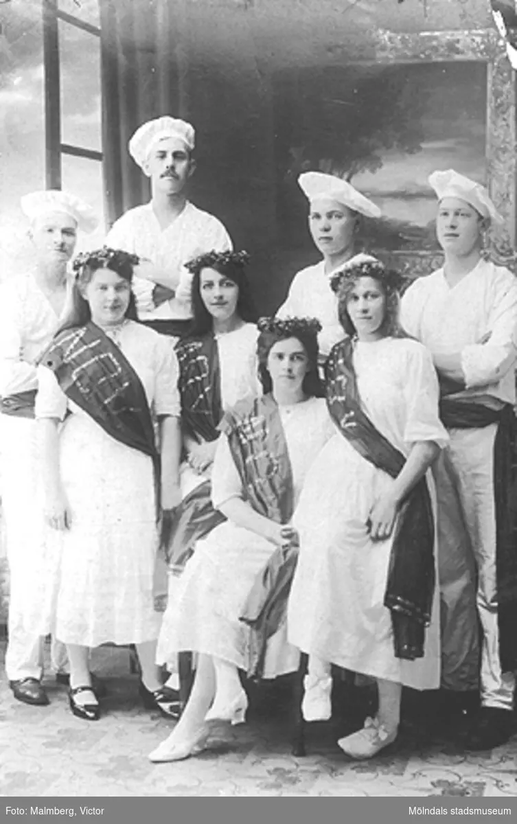 Foto från V. Malmbergs ateljé. Bild som postcard. Motiv: någon examensklass i början av 1900-talet. Flickorna bär banderoll med texten NOV.