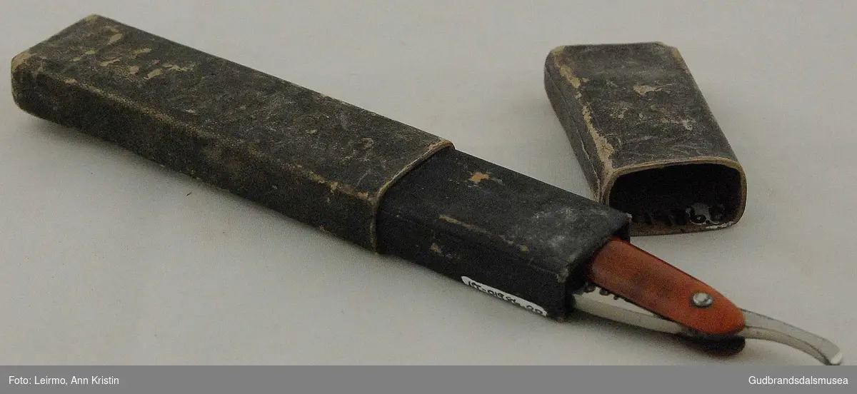 Sammenleggbar barberkniv, med svart og brunt skaft, har tilhørende etui.