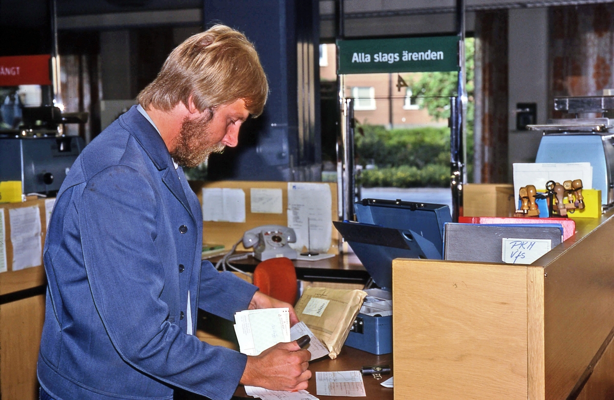 Mikael Mattsson räknar avier i kassan på  postkontoret  på Tuna torg i Vallentuna. Grå telefon med nummerskiva, ställ med stämplar, en våg, över luckan en skylt  Alla slags ärenden.