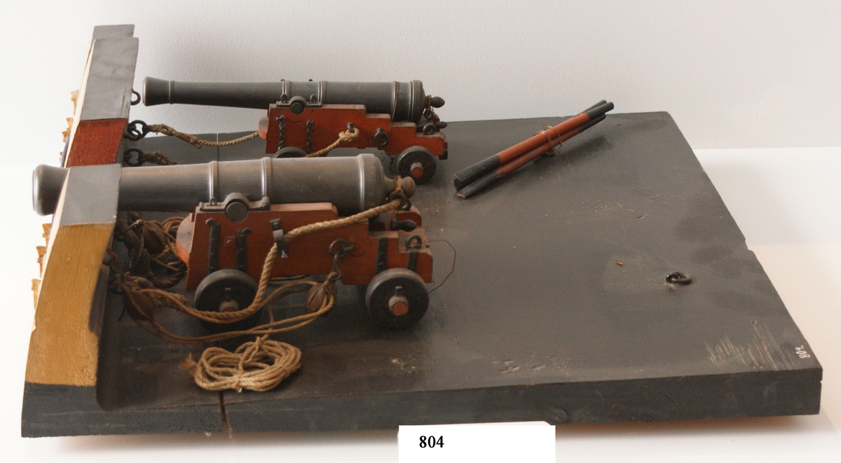 Kanonmodell: 24-pundig styckelåda av engelsk modell, med kanon. Modell av trä. Lavetten rödmålad, kanonen svart. Tillbehör: 1 st brok, 1 st riktkil, 2 st spakar och 2 st taljor. Lavetten L = 510 mm B = 480 H = 140 mm.