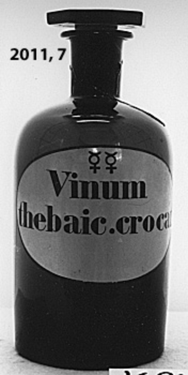 Flaska av brunt glas för medicin. Den är cylindrisk med glaspropp, försedd med plant, 8-kantigt huvud. På sidan är blå, ovalformad etikett med kant i svart målad. Märkning i svart: (se bild) Vinum thebaic. crocat.