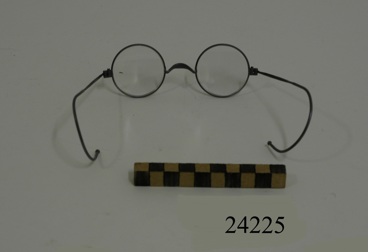 Glasögon med runda glas (D = 40 mm) infattade i runda bågar av grå metall. Skalmarna böjes bakom öronen. Glasögonen förvaras i grått plåtfodral med lock, MM 24226.