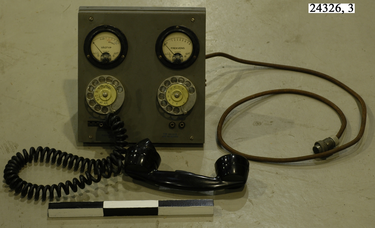 Grå-grön telefonapparat med två fingerskivor på framsidan. Ovanför fingerskivorna sitter två mätare, den vänstra för vågtyp den högra för frekvens. Till apparaten finns en anslutningskabel och en telefonlur av svart bakelit. På apparatens baksida finns skrivet med blått tusch: "MA 645, Tillhör, 800W-Sänd, Ras-720". Tillverkningsinformation på framsidan: TYP MA-645, APP. NR. 603152.