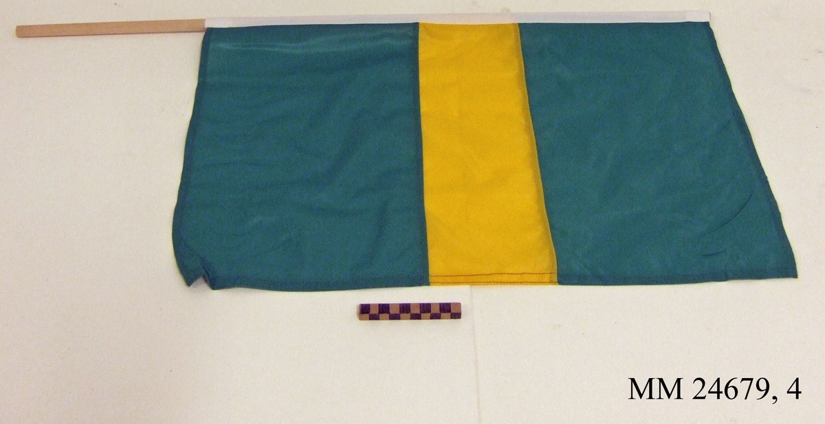 Manöverflagga på stång av trä. Flagga av grön nylon med gul hotisontal rand i mitten. Vitt lik trätt över rundstaven och fäst med häftklammer. Flaggan ligger i påse tillsammans med fem andra flaggor, sk. manöverflaggställ.