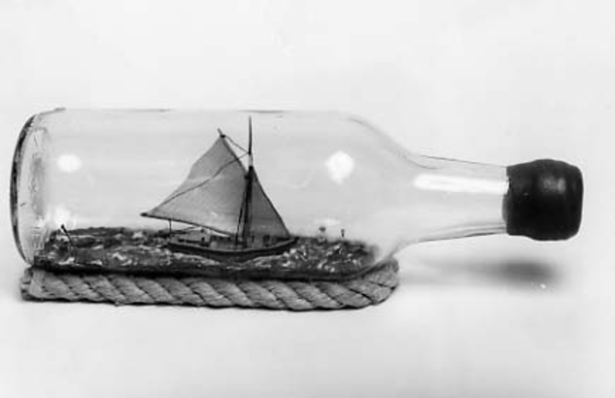 Flaskskeppsmodell av sandkil under segel med jolle på släp.
Flaskan vilar på skrå av otjärat rep.