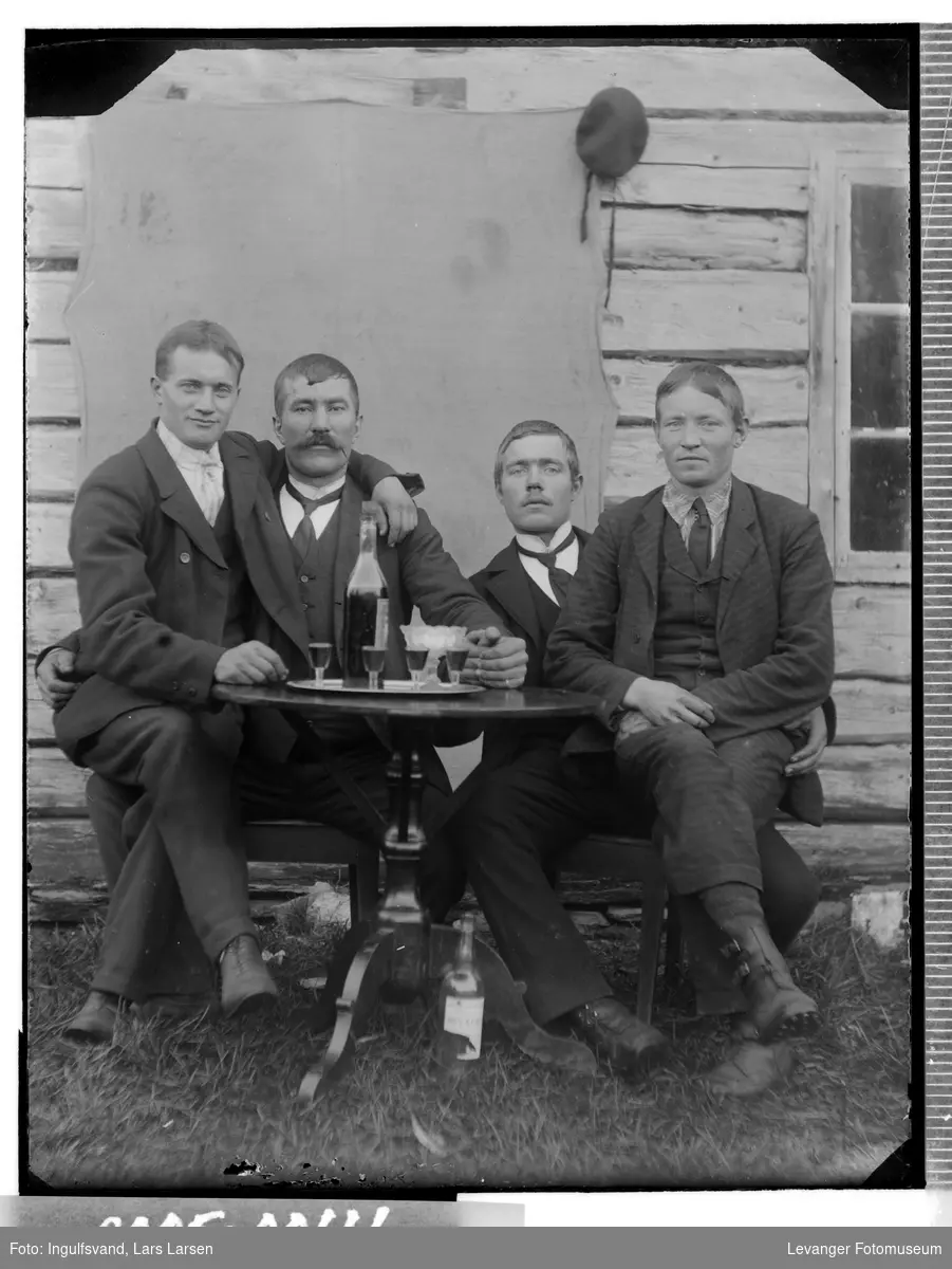 Gruppebilde av fire menn i festlig lag ved et bord med flasker og glass.