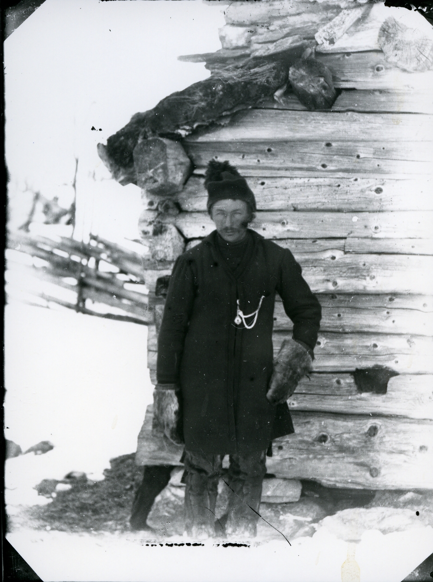 Trolig samisk mann, med skinnvotter, skinnbukse og lue med dusk. Avbildet foran laftet tømmerhus, vinterstid, med skigard i bakgrunnen. 
