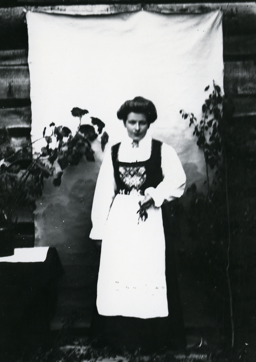 Kvinne kledd i nasjonaldrakt, avbildet i helfigur foran lerret på husvegg