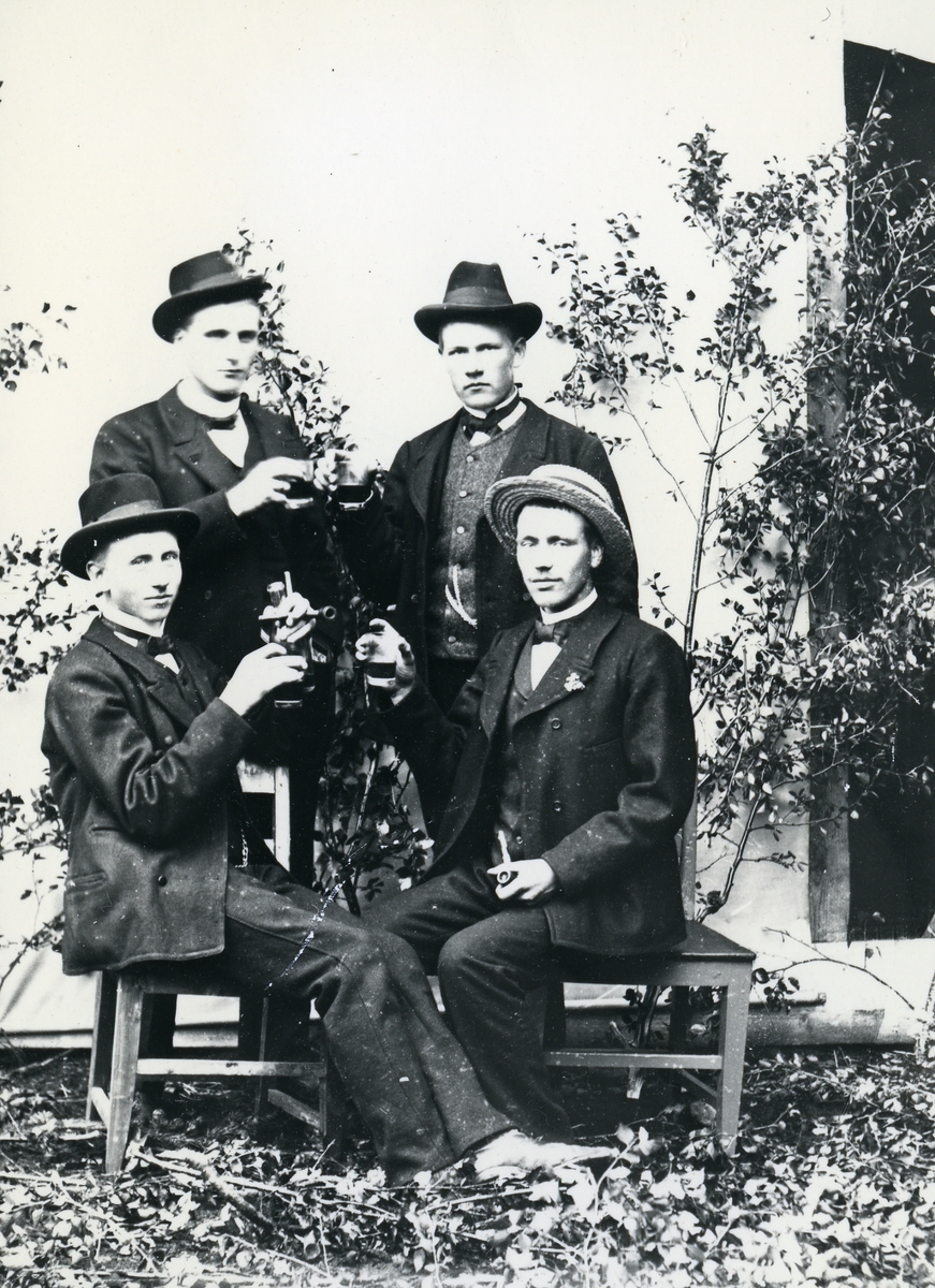 Fire menn kledd i dress og hatt, med glass og piper. Bjørkekvister og lerret som bakgrunn