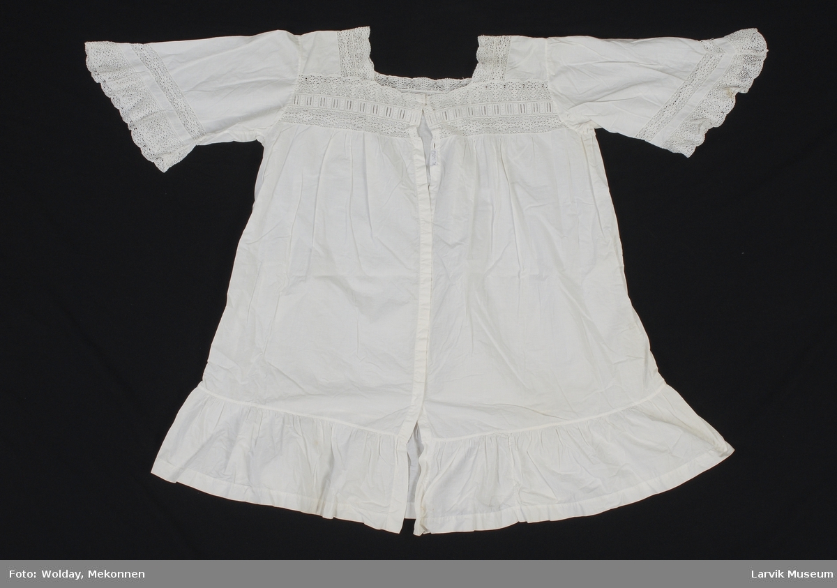 Form: Vid, kort "kjole" med knepping , maskinheklet blonde i halslinning og nederst på ermer. Perlemors knapper.
