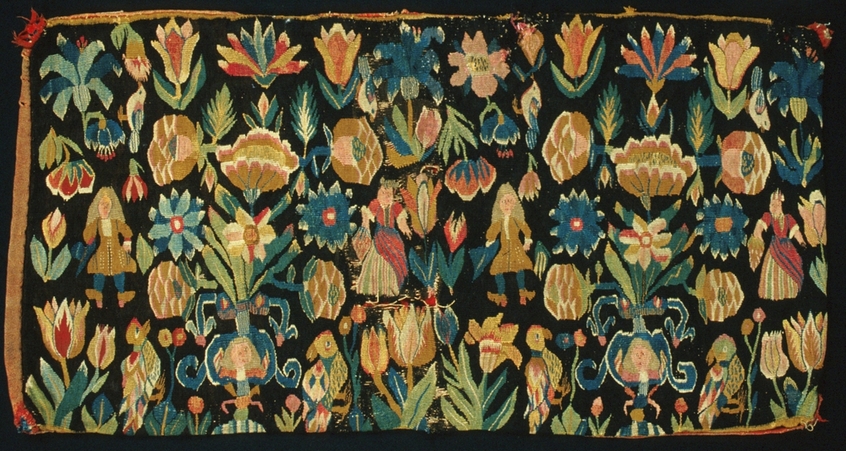 Dyna vävd i flamsk. En "blomsterdyna" med motiv av spridda blommor och fåglar, två urnor med människofigurer på, två kvinnofigurer i randiga kjolar och förkläden med frukter i händerna och två män i 1700-talsdräkter med varsin trekantig hatt med band i händerna. Brunsvart botten, mångfärgat mönster. Gul bård i kanten upp- och nedtill. Ursprungligen vadmalsklädda, röda kulor i hörnen.
Varp i s-tvinnat 2-trådigt oblekt lingarn, 4,5-6 trådar/cm. Inslag i 1-trådigt ullgarn, två trådar tillsammans. Invävt människohår i figurerna på urnorna och i mansfigurernas peruker. Guld- eller silvertråd (oxiderad) i mittkvinnofigurens huvudbonad och på bröstet, samt i bandet på mannens hatt. Vävnaden är av äldre typ och tekniken är nära besläktad med gobelinvävning. På baksidan kan man se sammanfogningar i skånsk rölakansnärjning, dvs snärjning av inslagstrådarna åt båda håll mellan varptrådarna, vilket ger en rygg/upphöjning på baksidan. Dessutom förekommer också enkel sammanfogning, dvs vändning runt en varptråd växelvis från vänster och höger, på flera ställen och i hela sidobården. Traditionell trestegstandning förekommer bara på ett ställe på hela dynan. 
Baksida av röd ylleväv i tuskaft. Varp och inslag av 1-trådigt s-spunnet ullgarn. Varptäthet: 17 trådar/cm, inslagstäthet: 10 trådar/cm. Spår av fjäder inuti. Märkt med tyglapp på baksidan med texten: "N° 200 a  Skytts h-d."