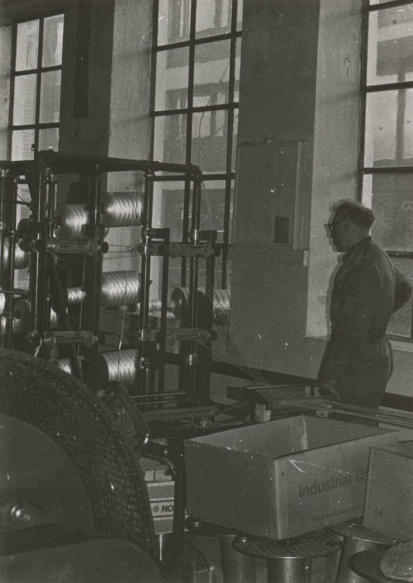 Interiørbilde fra maskinhallen ved Spilkevigs Snøre- Not- & Garnfabrik. En mann står å betjener maskinene.
