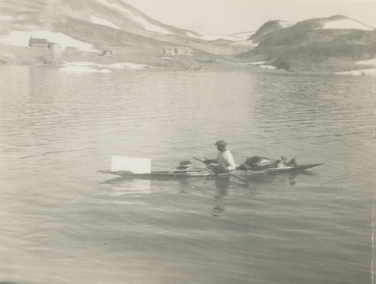 En mann fra Grønland er ute å padler i en kajakk med diverse utstyr ombord. Bebyggelse på land i bakgrunnen