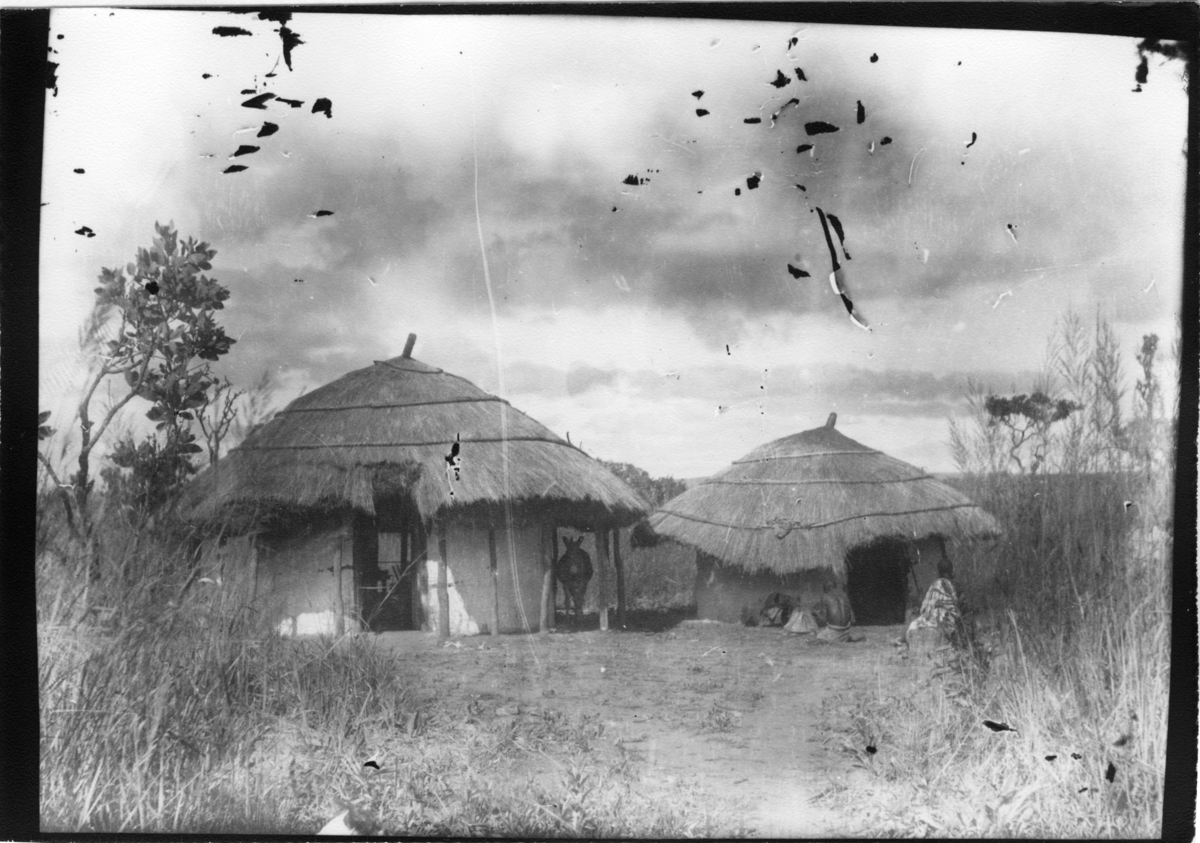 'Diverse fotografier från bl.a. dåvarande Nordrhodesia, nu Zambia, tagna av Konsul Magnus Leijer. ::  :: 2 cirkelformade enkla byggnader med halmtak, under det ena taket står en åsna. Framför den andra byggnaden sitter 2 människor.'