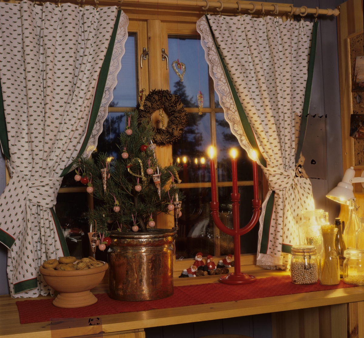 Et julepyntet vindu med gardiner, lys, kaker og et lite juletre. Illustrasjonsbilde fra Bonytt 1986.