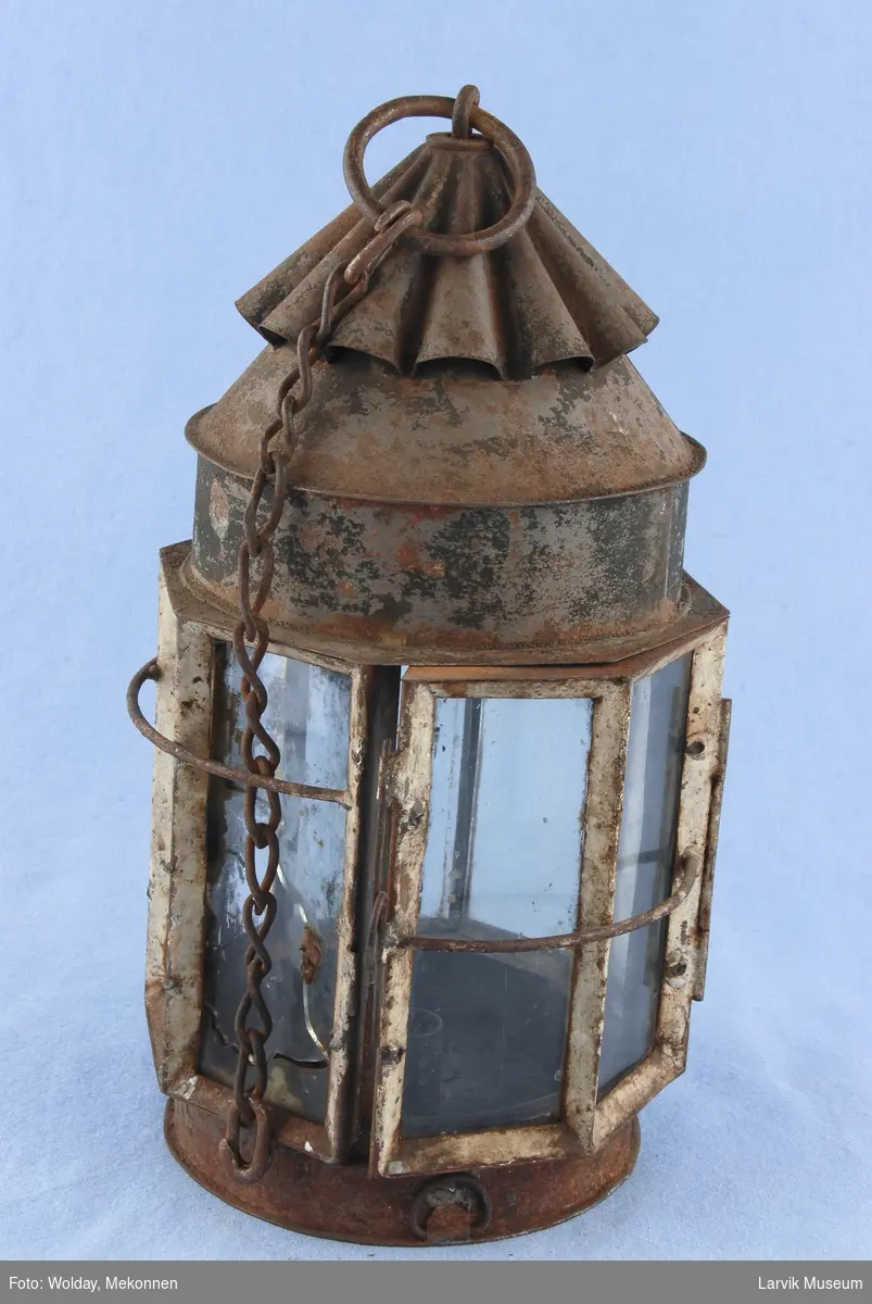 Sylinderformet lampe med bølgeformet toppstykke med ring for oppheng. 8 glassruter hvorav to utgjør dør.
