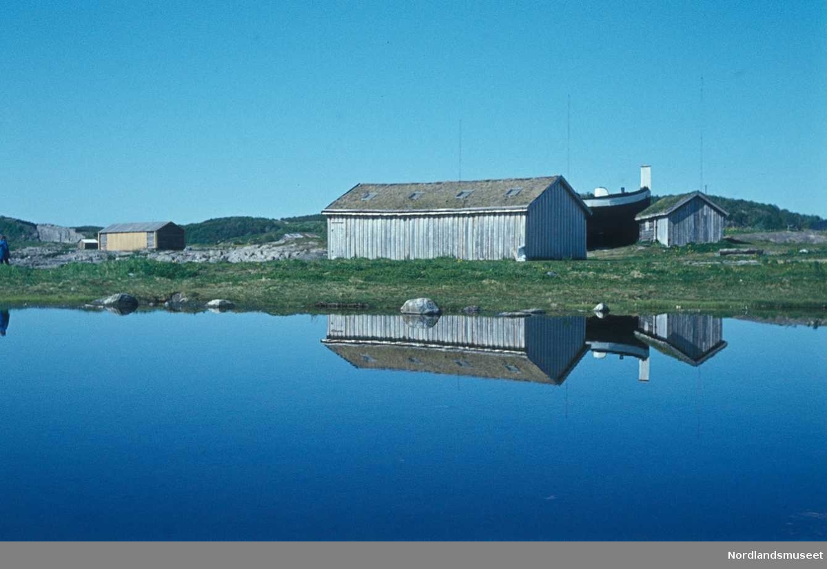 Dias fra Bodøsjøen frilufts museum, vi ser et umalt båthus med stående panel og torvtak, og ved siden av en mindre bu, bak sees jekta Anna Karolina delvis skjult av båthuset, forran ligg er havet speilblankt og det er gjenskinn fra husene, i kanten sees også en annen bygning, bak er det blå, klar, himmel.