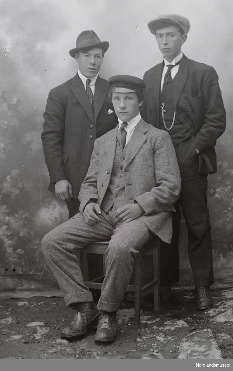 Portrett. Tre menn. En sitter på en stol, de andre står bak. De har alle på seg dress og slips med en hatt eller lue på hodet.