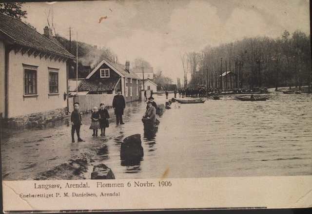 Flommen i Barbuelva 6. Novbr. 1906.  Elven i h. side. Barn og voksne på veien tv. og sitter på stabbesteinene. Prammer og mennesker i bakgrunnen. Veien er Vestlandske hovedvei.
