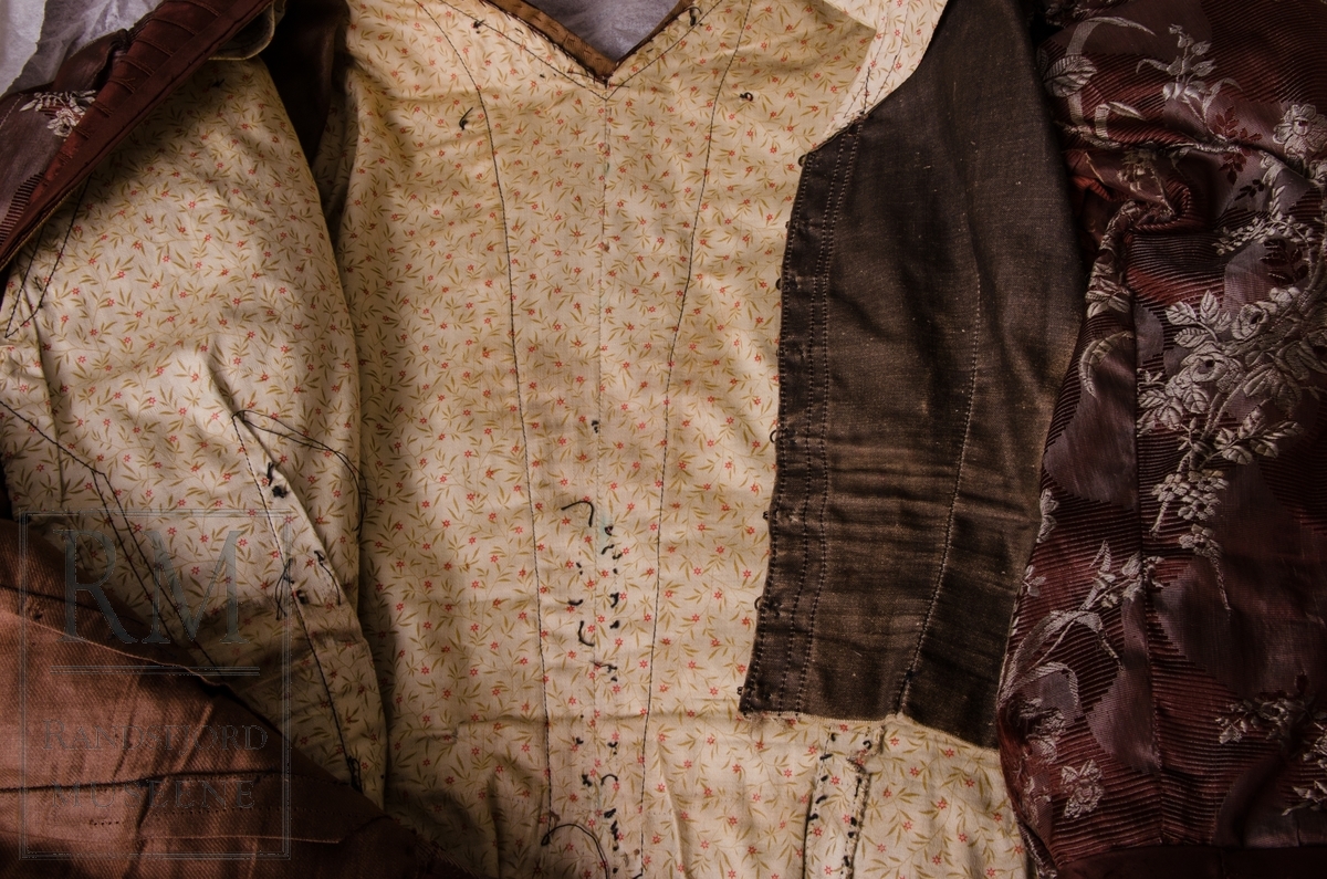 Todelt. Skjørt og kjoleliv i brun/grå silkedamask med brun silkebesetning. Brunt silkebelte. Kjolelivets bol er foret med lys bomullssatin med trykt blomstermønster. Kjolen er blitt omsydd rundt forrige århundreskifte.