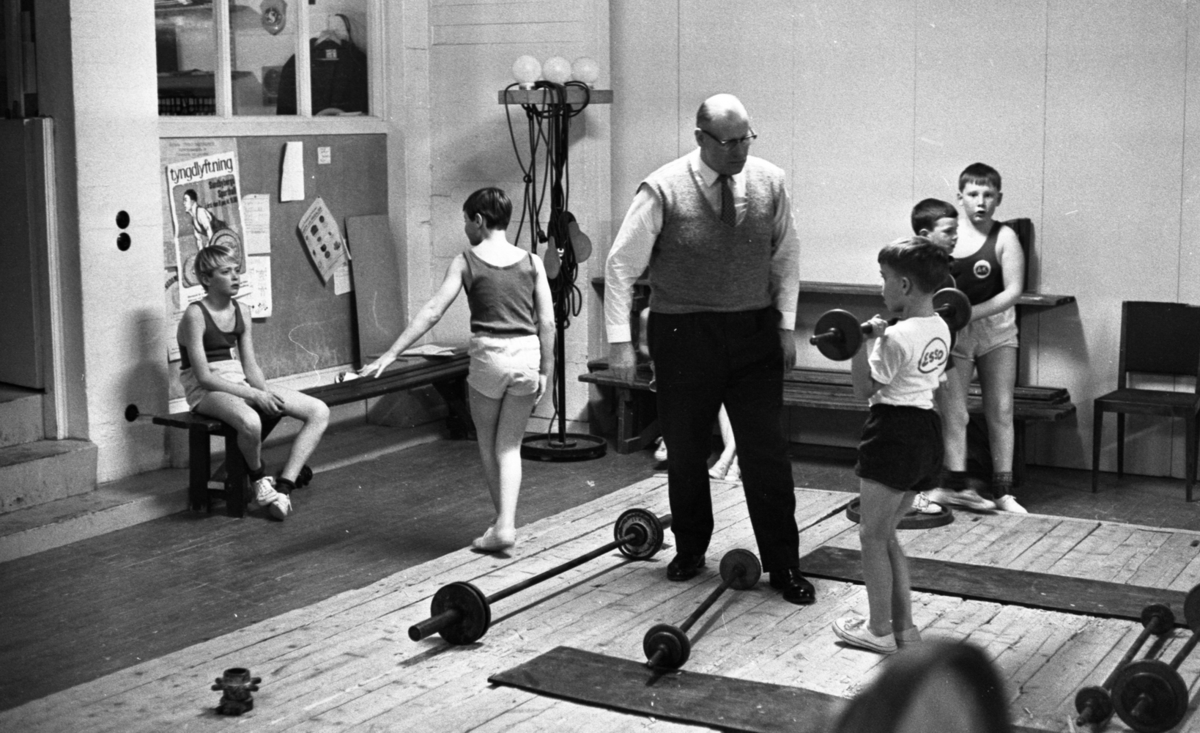 Tyngdlyftarreportage 31 januari 1966 

En tränare, en äldre man, klädd i skjorta, slips, väst, byxor och skor instruerar en liten pojke i åttaårsåldern som har lyft upp en skivstång i axelhöjd. Den lille pojken är klädd i T-shirt, shorts och träningsskor. Fyra andra skivstänger ligger på golvet. I bakgrunden sitter en pojke på en bänk och tre andra pojkar står upp. Två avlånga träningsmattor ligger på golvet.