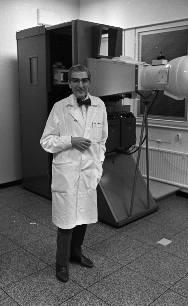 Lungkliniken, Nappivalen, Grekiskt handarbete 11 december 1967

En läkare i vit läkarrock, vit skjorta, svart fluga, mörkgrå byxor och svarta skor. En stor röntgenapparat står bakom honom (medicinsk utrustning). Ett fönster syns i bakgrunden även.