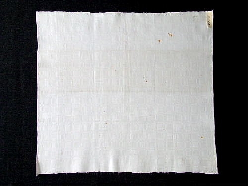 Vit servetten i bomull från Fårö. Vävd i sk "Fattigmansdräll", 4-skaft. Mönstret är likrutigt över servetten, med undantag av en bård runt kanten med mindre rutmönster.