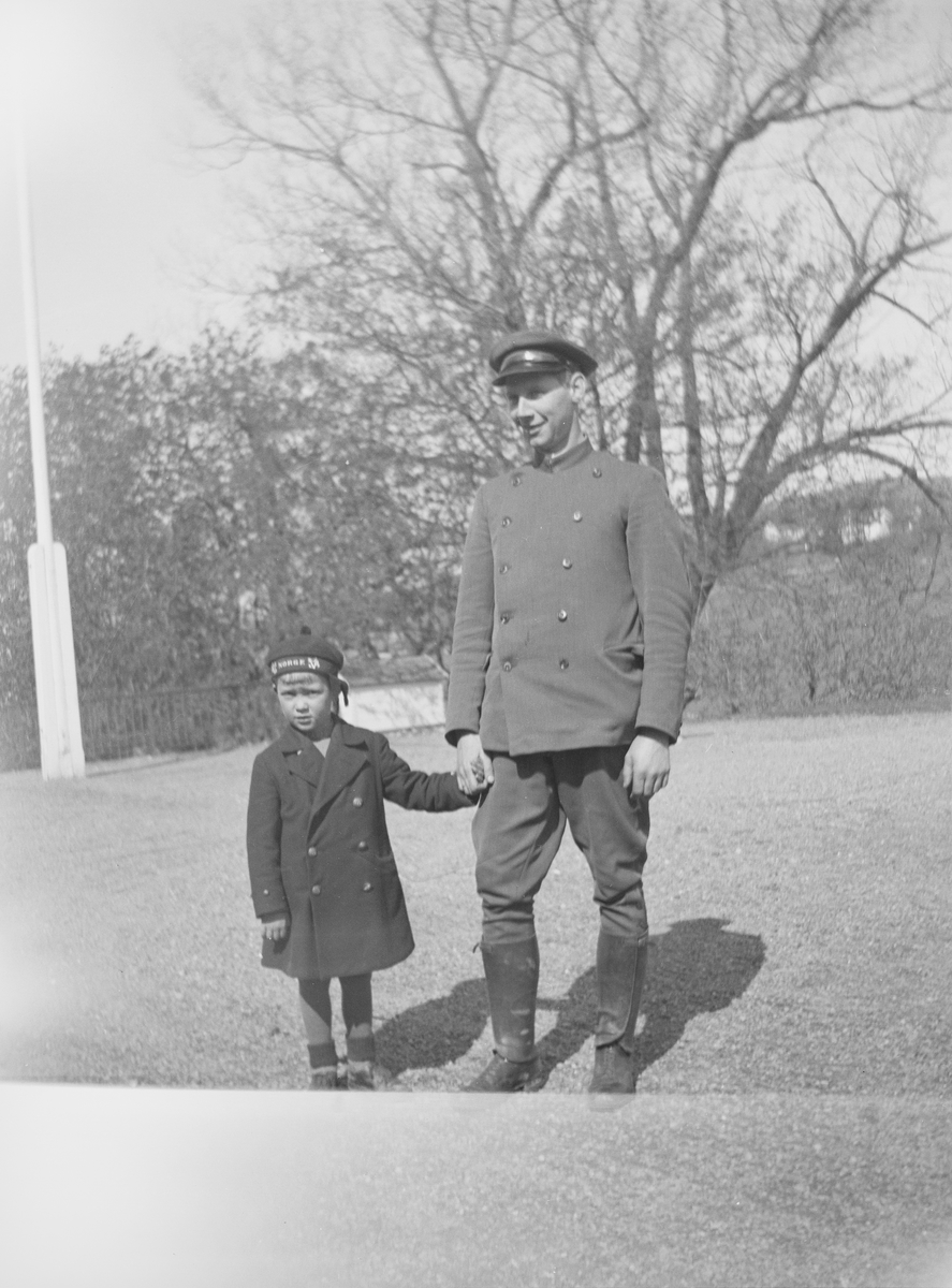 En mann i en sivil uniform holder en gutt i hånden. De står på gårdsplassen på Linderud Gård. Det er vår.