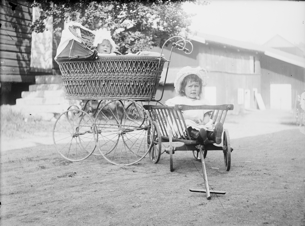 To barn på et gårdstun; den ene sitter i en tralle av tre og det minste i en barnevogn.