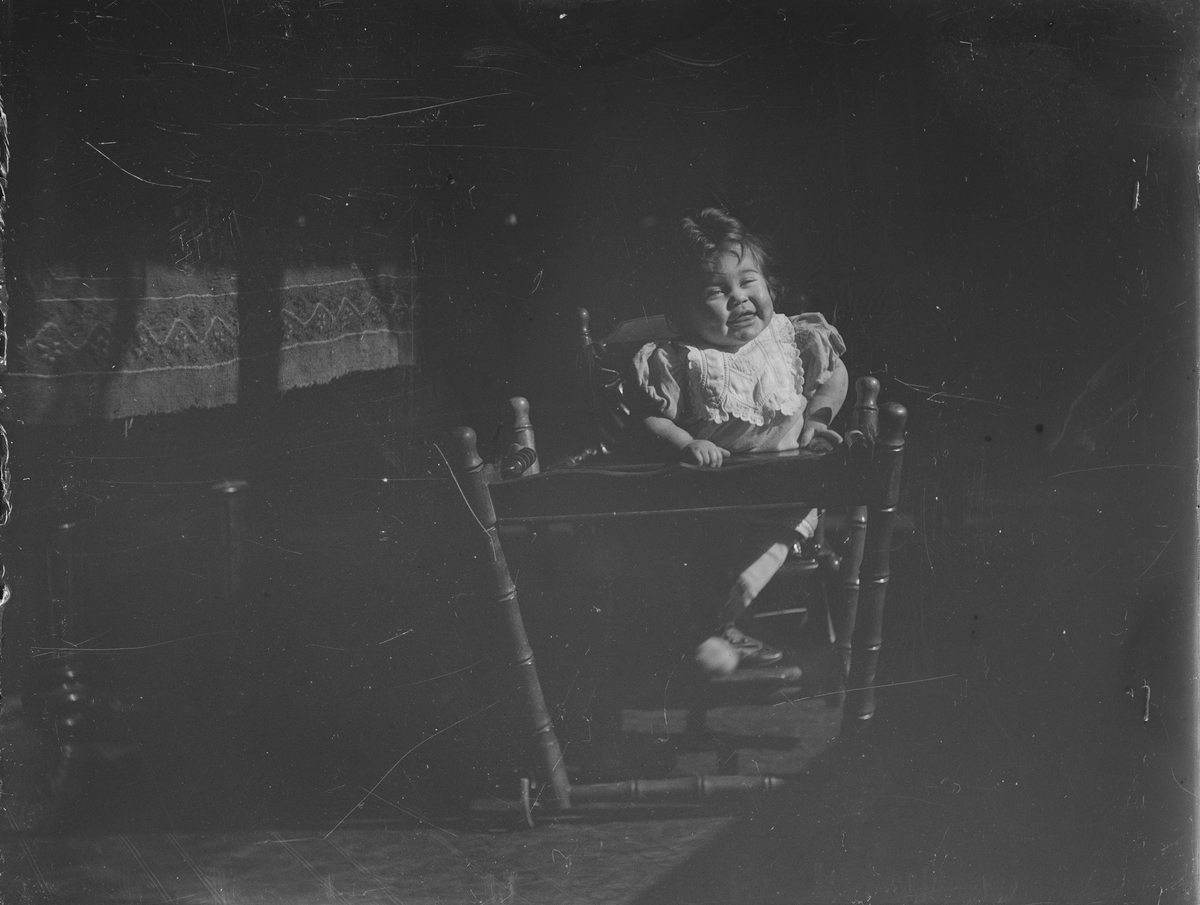Et lite barn sitter smilende i et barnemøbel; en lav barnestol med bord. Det ser ut som det er hjul montert på bordbena.