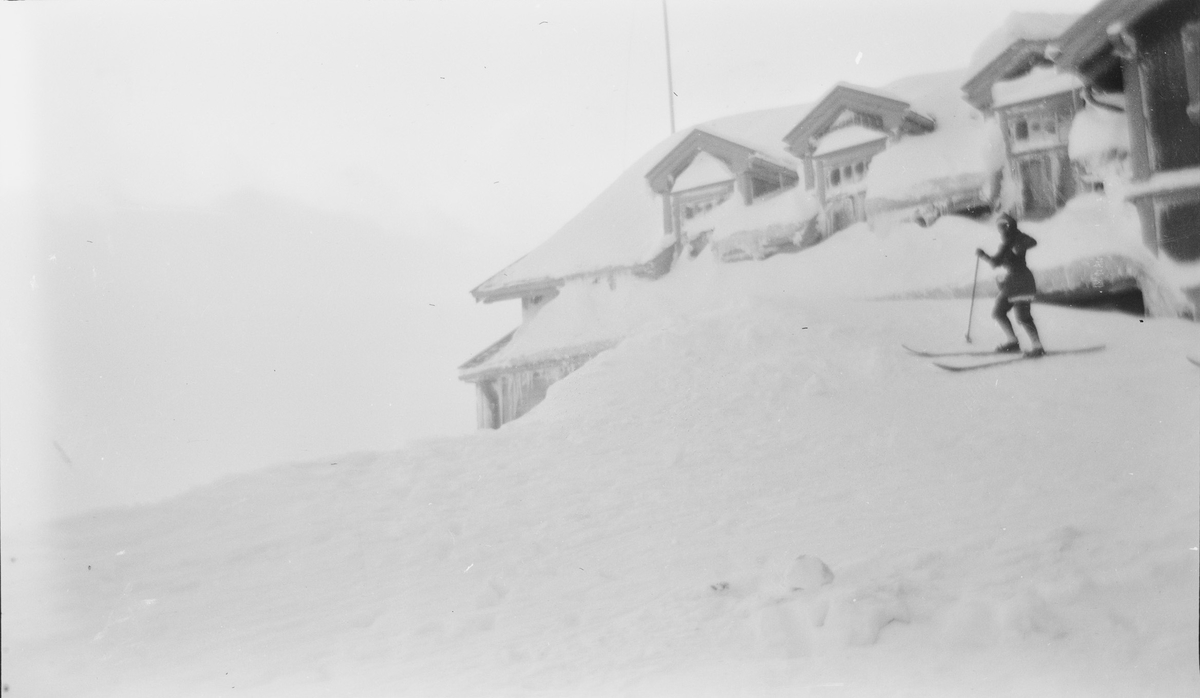 En person, på ski, står nesten oppe på taket av Haugastøl stasjon og turisthotel.