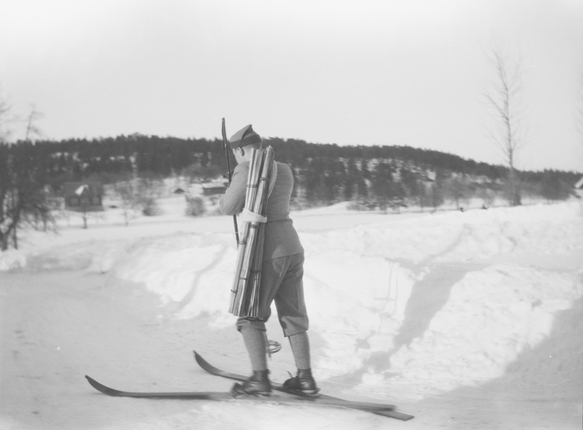 En skiløper med ryggen til fotografen ser ut til å være på vei bort fra gårdsplassen på Linderud Gård. På ryggen har han en bunt med fakler.