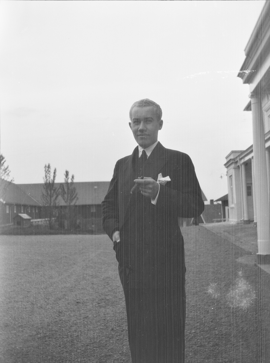 Iacob Ihlen Mathiesen står på gårdsplassen utenfor hovedhuset på Linderud Gård med en sigar i hånden. I bakgrunnen sees driftsbygninger tilhørende gården.